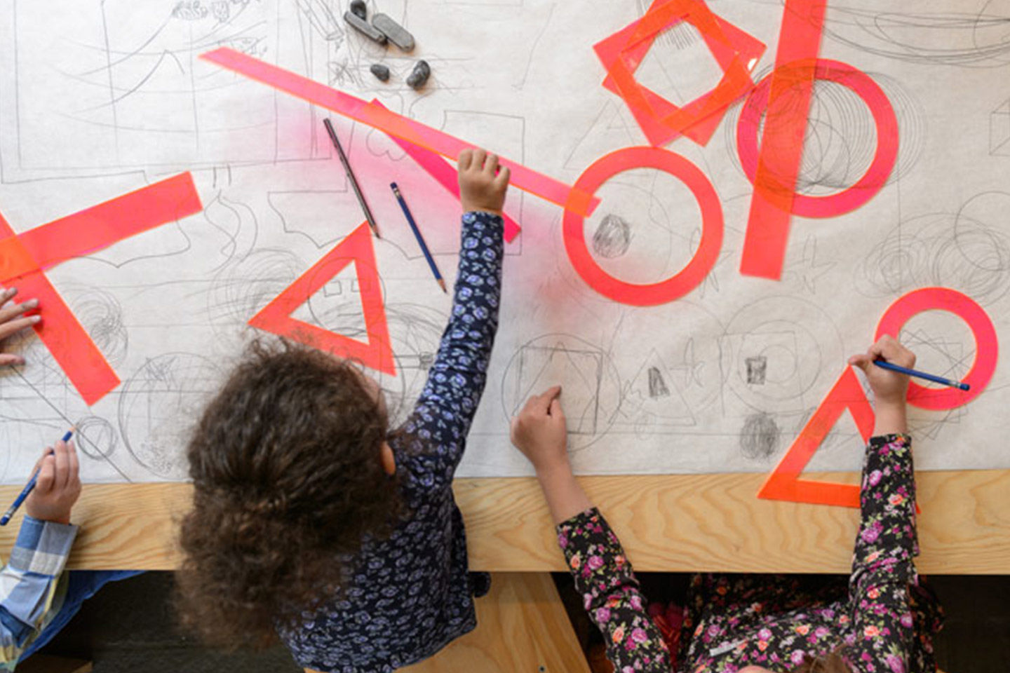 Overhead view of three small children seated at a wooden table draped in one long sheet of white paper covered in pencil drawings; the children draw and interact with assorted neon orange and pink geometric stencils